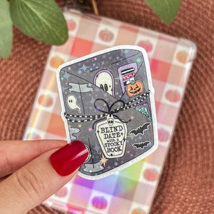 Peut inclure: Un autocollant holographique sur le thème d'un livre effrayant. L'autocollant représente un livre gris avec un fantôme, une citrouille et des chauves-souris. Le texte dit "Blind Date with a Spooky Book". L'autocollant est tenu par une main avec du vernis à ongles rouge.