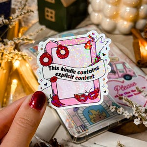 Op de afbeelding: Een roze en witte sticker met glitter en een rode omtrek. De sticker heeft een cartoon afbeelding van een Kindle met vuur en een masker. De tekst op de sticker luidt "This kindle contains explicit content".