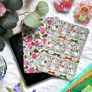 Peut inclure: Liseuse Kindle avec deux étuis à motifs. L'un présente des fraises et des fleurs, l'autre des oies et des nœuds. Des épingles décoratives d'un panier de pique-nique et d'un ours en chapeau de fleur sont également visibles.
