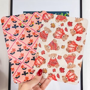 May include: Two decorative cards with different patterns. One card features a pink floral design with orange flowers and black accents. The other card has a pattern of teddy bears wearing red sweaters and autumn leaves.