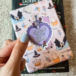 Könnte beinhalten: Ein violetter herzförmiger Aufkleber mit glitzernder Oberfläche. Der Aufkleber zeigt ein Schwert und den Text "violent little thing" in einer gotischen Schrift.