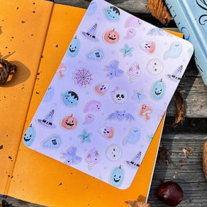 May include: A rectangular, light purple notebook featuring Halloween-themed illustrations. The design includes pumpkins, ghosts, witch hats, bats, and tombstones. The notebook is open on top of an orange book, with a candle and book in the background.