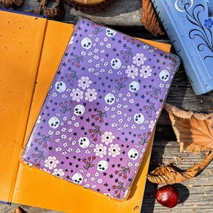 Peut inclure: Une coque de tablette transparente avec un motif dégradé violet orné de crânes blancs, de fleurs et d'étoiles. L'étui repose sur un livre ouvert aux pages orange. Une bougie dans un pot en verre et un livre bleu sont également visibles.