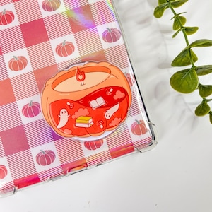 May include: A clear phone case with a red and pink gingham pattern and pumpkin illustrations. A pumpkin-shaped sticker with a candle, ghosts, books, and mushrooms is attached to the case. A sprig of green leaves is in the background.