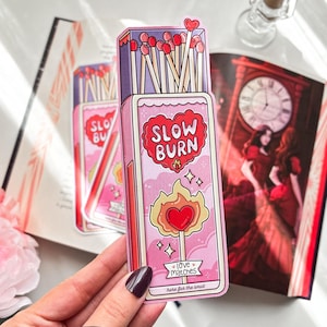 Peut inclure: Autocollant rose en forme de boîte d'allumettes avec les mots "SLOW BURN" dans un cœur rouge. L'autocollant présente une flamme avec un cœur et le texte "love matches" et "here for the smut".