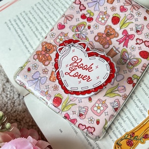 Könnte beinhalten: Eine transparente Handyhülle mit einem rosa Muster aus Teddybären, Erdbeeren, Schleifen und Schmetterlingen. Ein herzförmiger Pop-Socket in Rot und Weiß mit der Aufschrift "Book Lover" ist angebracht. Die Hülle liegt auf einem offenen Buch.