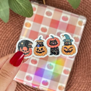 Puede incluir: Un conjunto de cuatro pegatinas con temática de Halloween con diseños de calabazas y gatos. Las pegatinas tienen un acabado transparente y brillante y están sujetas por una mano con esmalte de uñas rojo. El fondo es un patrón de cuadros con ilustraciones de calabazas.