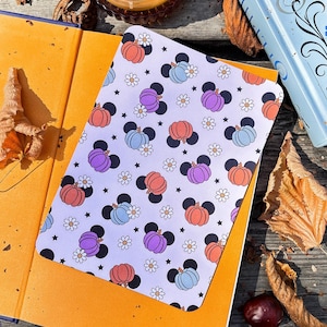 May include: A notebook with a light purple cover featuring a pattern of pumpkins in orange, purple, and blue with black mouse ears, white daisies, and black stars. The notebook is open on top of an orange book, with a candle and leaves in the background.