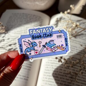 May include: A purple and blue sticker with a fantasy book club theme. It features a small dragon, a sword, a key, a cup of tea, and a stack of books. The sticker says "Fantasy Book Club" and "Admit One" on either side.