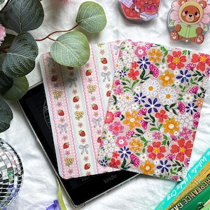 Peut inclure: Un lecteur Kindle avec deux étuis décoratifs à motifs floraux et fraises. L'image comprend des épingles décoratives, des livres et des fleurs, créant une esthétique fantaisiste et féminine.