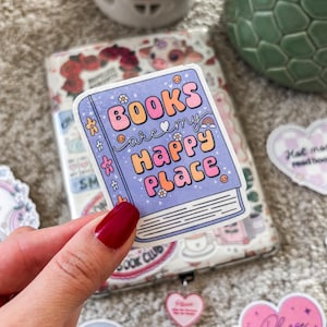 Puede incluir: Una pegatina morada y azul con el texto "BOOKS are my HAPPY Place" en letras de colores. La pegatina tiene forma de libro con una columna vertebral blanca y un arcoíris.