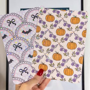 Może przedstawiać: Dwie karty dekoracyjne o tematyce Halloween i jesieni. Jedna karta przedstawia wzór kokardek i nietoperzy, druga dynie i fioletowe kwiaty. Obie karty mają błyszczące, opalizujące wykończenie.