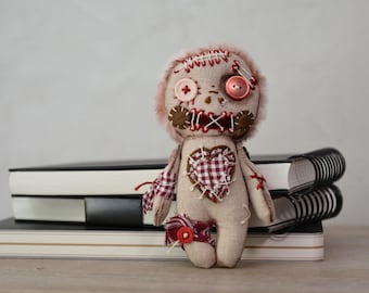 May include: A handmade plush doll with a stitched face, button eyes, and a heart-shaped patch. The doll has a fuzzy head and is dressed in beige fabric with red and white accents. It is sitting on a stack of notebooks.