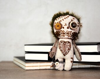 May include: A handmade fabric doll with button eyes, stitched mouth, and a heart-shaped patch. The doll has brown and beige accents, with a green fur-like hair. It is standing in front of a stack of books.