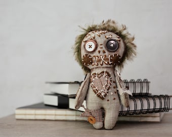 May include: A handmade fabric doll with button eyes and stitched details. The doll has a heart-shaped patch on its chest and is adorned with brown and beige fabrics. It is posed in front of a stack of books and a spiral notebook.