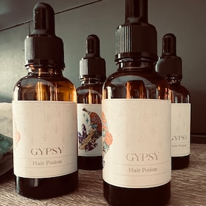 May include: Four brown glass bottles with black droppers. Each bottle has a white label with the word "GYPSY" and "Hair Potion" printed on it. The bottles are arranged on a wooden surface.