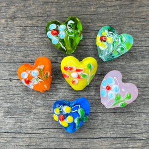 May include: Six glass heart-shaped beads with floral designs. The beads are in various colors, including green, blue, yellow, orange, and pink.