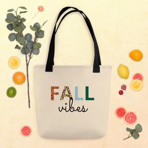 Puede incluir: Una bolsa de tela beige con asas negras. La bolsa tiene un diseño con la palabra "FALL" en letras de colores con patrones y las palabras "vibes" en escritura negra debajo.