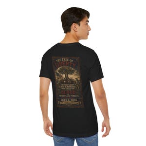 May include: A black short-sleeved t-shirt with a rectangular graphic on the back. The design features a tree, an American flag, and soldiers with the text: THE TREE OF LIBERTY MUST BE REFRESHED FROM TIME TO TIME WITH THE BLOOD OF PATRIOTS AND TYRANTS. JULY 4, 2026 250 YEARS OF FREEDOM.