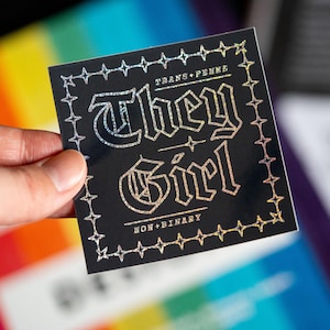 May include: A black sticker with a holographic finish features the text "They Girl" in a stylized font. The text "Trans + Femme" appears above the main text and "Non-Binary" appears below.