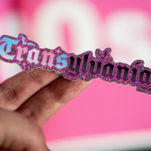 Puede incluir: Una pegatina holográfica con la palabra "Transylvania" en una fuente gótica. El texto está delineado en negro y relleno con degradados azules y rosas. La pegatina tiene un borde brillante y está sujeta por una mano.
