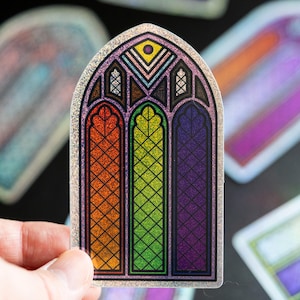 Può includere: Un adesivo con un disegno di vetrata color arcobaleno. L'adesivo ha un bordo argentato glitterato. La finestra è divisa in tre sezioni, ognuna con un colore diverso. L'adesivo è tenuto su uno sfondo scuro.