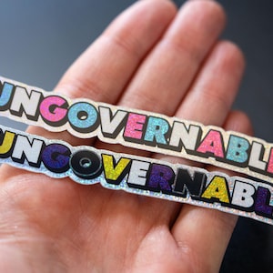 May include: Two silver glitter stickers with the word "UNGOVERNABLE" in black letters with colourful accents. The top sticker has pink, blue, and white accents. The bottom sticker has yellow, purple, and white accents.