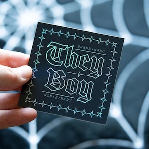 Może przedstawiać: Czarna naklejka z białą obwódką i holograficznym wykończeniem. Naklejka zawiera napis "They Boy" w stylu gotyckim. Na naklejce znajduje się również tekst "Trans-Masc" i "Non-Binary".