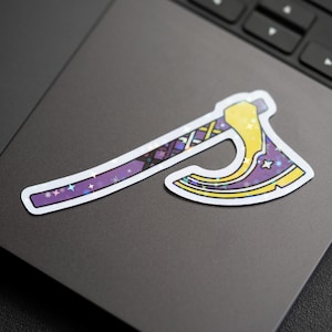 Op de afbeelding: Een sticker van een gestileerde bijl met een paarse steel en een gele kling. De steel heeft een geometrisch patroon en de kling heeft steraccenten. De sticker zit op een laptop.