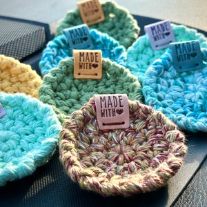 May include: A collection of seven crocheted coasters in various colors, including green, blue, yellow, and brown. Each coaster has a small leather tag attached with the text "Made with Love".