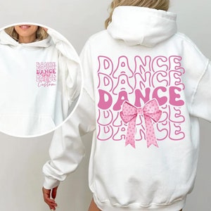 May include: White hoodie with a pink graphic of the word "DANCE" repeated multiple times with a pink bow in the centre. The hoodie has a drawstring hood.