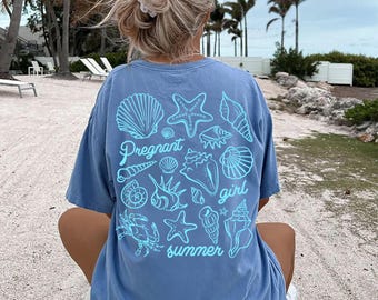 May include: A blue t-shirt with a light blue seashell and starfish design, and the words "Pregnant girl summer". The shirt is worn by a person sitting on a beach with palm trees in the background.