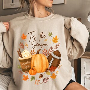 May include: Beige sweatshirt featuring a seasonal design. The graphic includes a pumpkin spice latte, pumpkins, a rugby ball, and autumn leaves. The text "Tis the Season" is written in a script font.