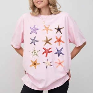 May include: A pink t-shirt with a graphic print of 12 colorful starfish. The starfish are arranged in a grid pattern.