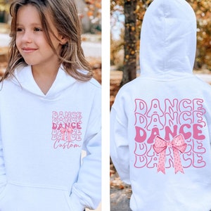 May include: White hoodie with a pink bow and the word "DANCE" repeated in a stacked design. The front of the hoodie has the word "DANCE" and "Custom" printed below a bow. The back of the hoodie has the word "DANCE" repeated.