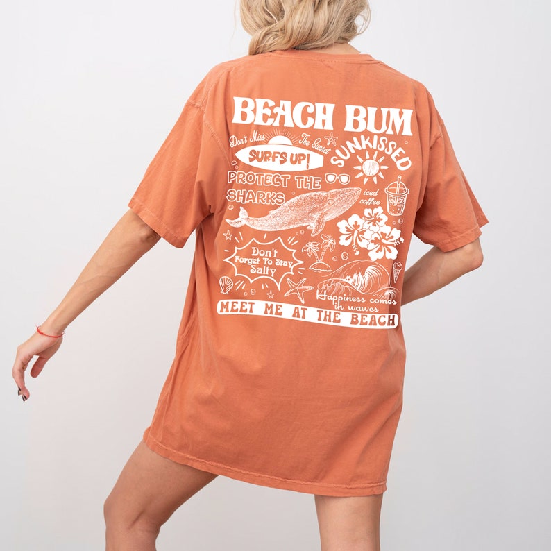May include: Burnt orange t-shirt featuring a beach-themed design on the back. The graphic includes the words "Beach Bum," "Surfs Up," and "Meet Me at the Beach," alongside illustrations of a whale, sunglasses, and palm trees.