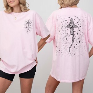 Puede incluir: Camisetas de color rosa claro con un dise&ntilde;o de tibur&oacute;n en blanco y negro. Una camiseta tiene un peque&ntilde;o gr&aacute;fico de tibur&oacute;n en la parte delantera, y la otra tiene un dise&ntilde;o de tibur&oacute;n m&aacute;s grande en la parte trasera, rodeado de estrellas y l&iacute;neas. Las camisetas son de gran tama&ntilde;o.