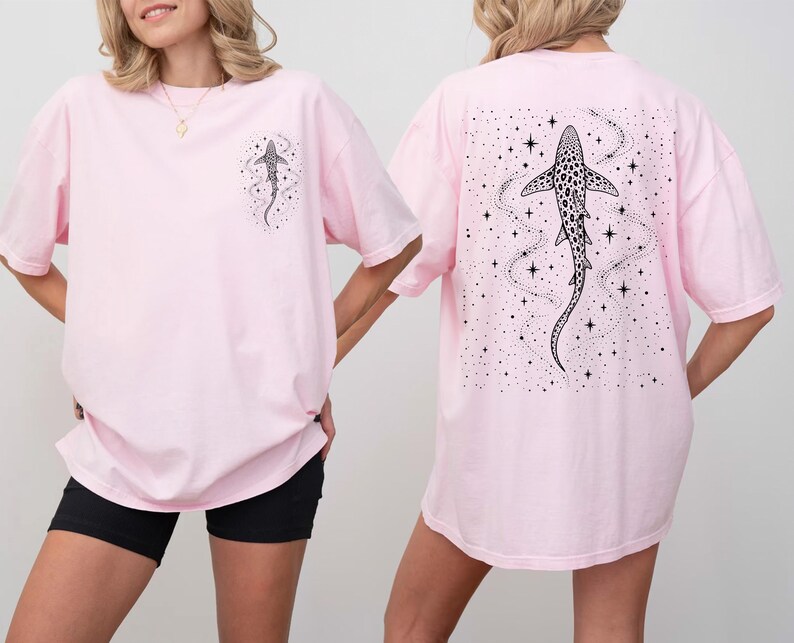 Puede incluir: Camisetas de color rosa claro con un dise&ntilde;o de tibur&oacute;n en blanco y negro. Una camiseta tiene un peque&ntilde;o gr&aacute;fico de tibur&oacute;n en la parte delantera, y la otra tiene un dise&ntilde;o de tibur&oacute;n m&aacute;s grande en la parte trasera, rodeado de estrellas y l&iacute;neas. Las camisetas son de gran tama&ntilde;o.