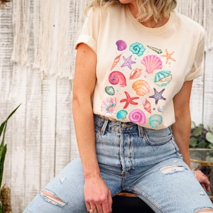 K&ouml;nnte beinhalten: Ein hellbeiges T-Shirt mit einem farbenfrohen Muschel-Design. Das Shirt zeigt eine Vielzahl von Muscheln in den Farben Rosa, Blau, Gr&uuml;n und Orange.