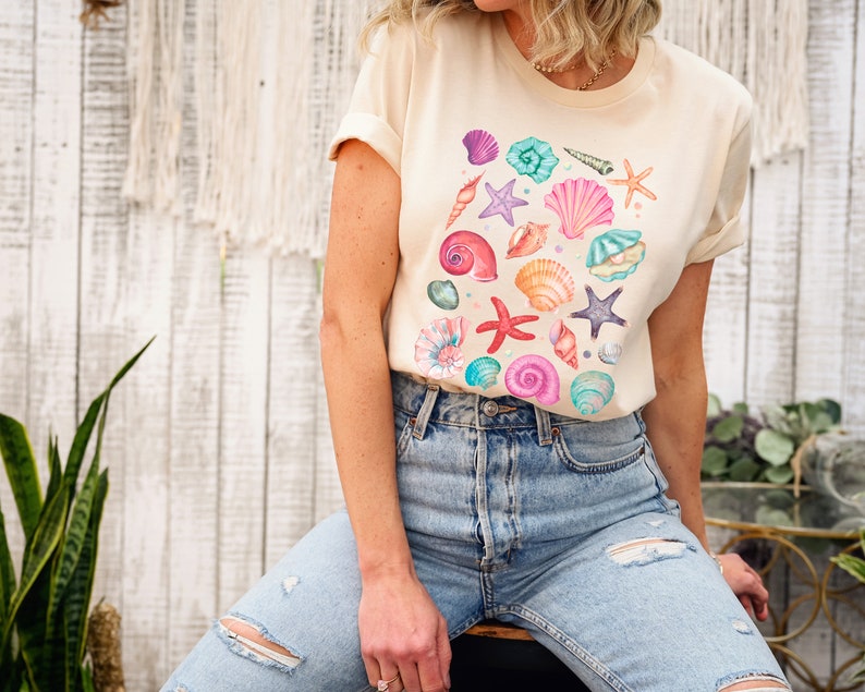 K&ouml;nnte beinhalten: Ein hellbeiges T-Shirt mit einem farbenfrohen Muschel-Design. Das Shirt zeigt eine Vielzahl von Muscheln in den Farben Rosa, Blau, Gr&uuml;n und Orange.