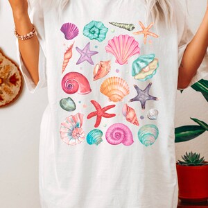 K&ouml;nnte beinhalten: Wei&szlig;es T-Shirt mit einem bunten Muster aus Muscheln und Seesternen. Das Design zeigt verschiedene Arten von Muscheln in den Farben Rosa, Lila, Blau und Orange sowie Seesterne in den Farben Lila und Rot.