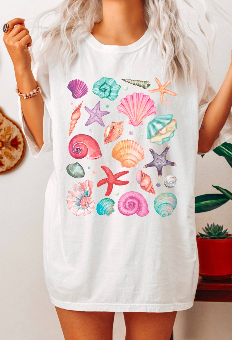 K&ouml;nnte beinhalten: Wei&szlig;es T-Shirt mit einem bunten Muster aus Muscheln und Seesternen. Das Design zeigt verschiedene Arten von Muscheln in den Farben Rosa, Lila, Blau und Orange sowie Seesterne in den Farben Lila und Rot.