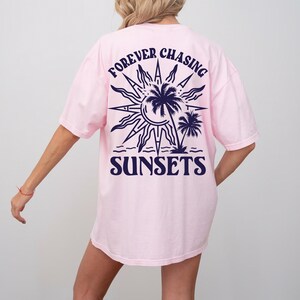 Puede incluir: Camiseta rosa de gran tama&ntilde;o con un gr&aacute;fico azul marino de un sol, palmeras y el texto "FOREVER CHASING SUNSETS".