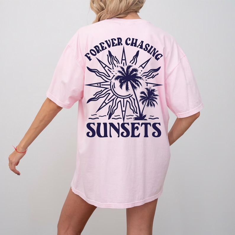 Puede incluir: Camiseta rosa de gran tama&ntilde;o con un gr&aacute;fico azul marino de un sol, palmeras y el texto "FOREVER CHASING SUNSETS".