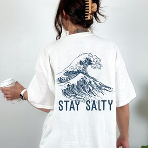 May include: White t-shirt with a blue wave graphic and the text "STAY SALTY" printed on the back.