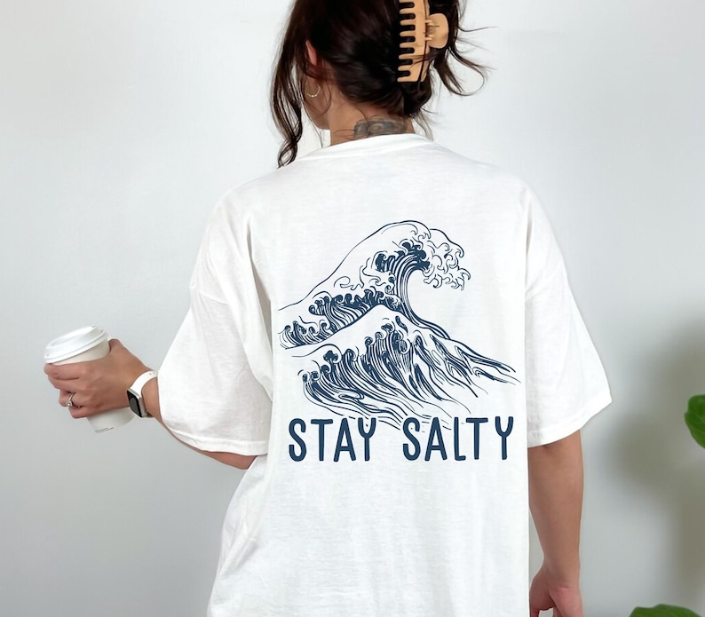 May include: White t-shirt with a blue wave graphic and the text "STAY SALTY" printed on the back.