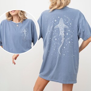 Puede incluir: Camiseta azul polvoriento con un dise&ntilde;o de tibur&oacute;n ballena blanco en la espalda y una versi&oacute;n m&aacute;s peque&ntilde;a en la parte delantera. El dise&ntilde;o incluye estrellas y un tema c&oacute;smico. La camiseta tiene mangas cortas y un corte holgado.