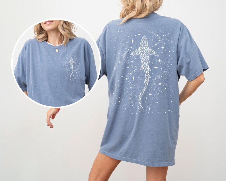 Puede incluir: Camiseta azul polvoriento con un dise&ntilde;o de tibur&oacute;n ballena blanco en la espalda y una versi&oacute;n m&aacute;s peque&ntilde;a en la parte delantera. El dise&ntilde;o incluye estrellas y un tema c&oacute;smico. La camiseta tiene mangas cortas y un corte holgado.