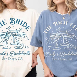 May include: Two light blue t-shirts with a white graphic design. The graphic features a group of hands holding up martini glasses with the text "The Bride" and "The Bach Club" on the shirts. The text "Emily's Bachelorette San Diego, CA" is also on the shirts.