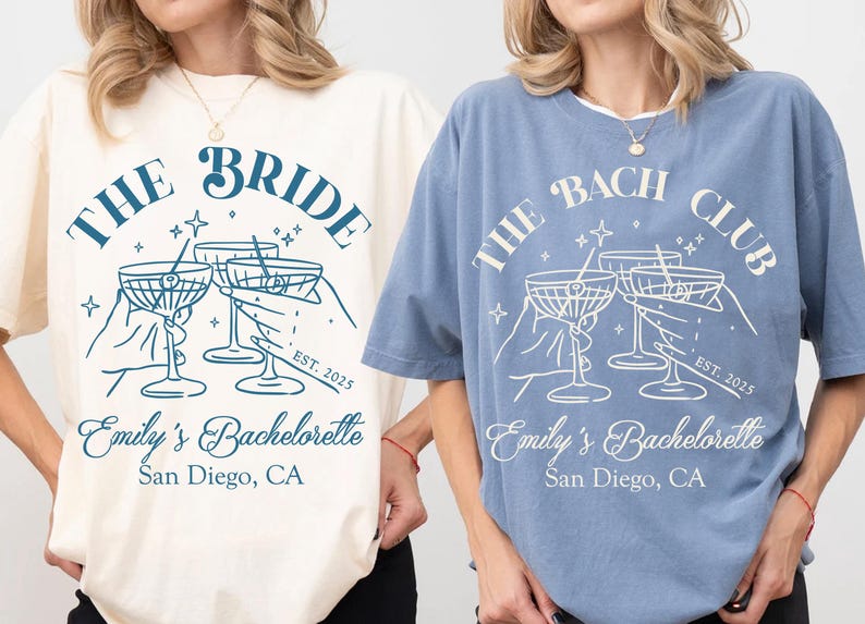 May include: Two light blue t-shirts with a white graphic design. The graphic features a group of hands holding up martini glasses with the text "The Bride" and "The Bach Club" on the shirts. The text "Emily's Bachelorette San Diego, CA" is also on the shirts.